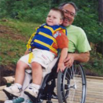 Older man in a wheelchair on a dock. He holds a young boy on his lap.