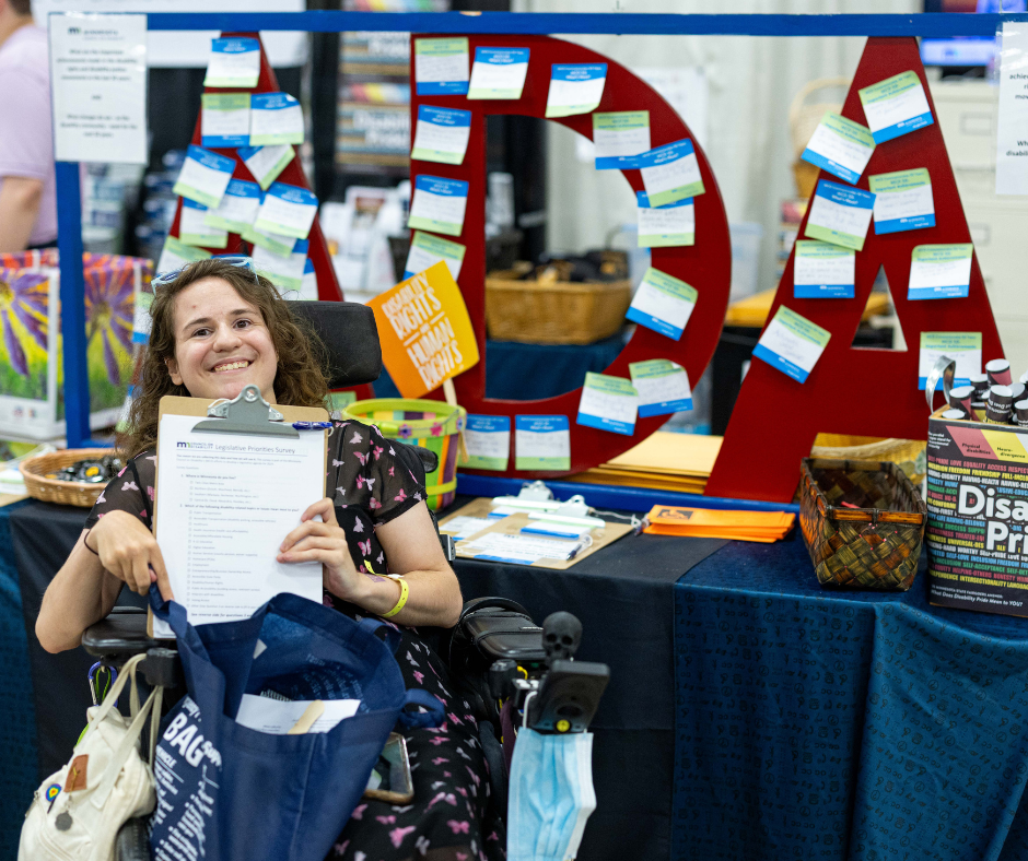 Woman who uses a wheelchair, holding a copy of the public policy survey and smiling. She is sitting in front of a table with a red ADA sign at the MCD State Fair booth.