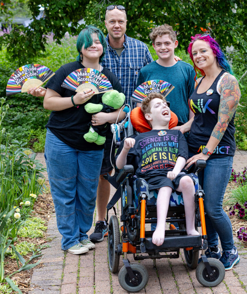 A group of people smiling and holding Pride-themed fans printed with the words, "Make Pride Accessible." One of the people uses a wheelchair.