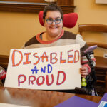 A woman using a wheelchair holds a sign that reads, "Disabled and Proud."