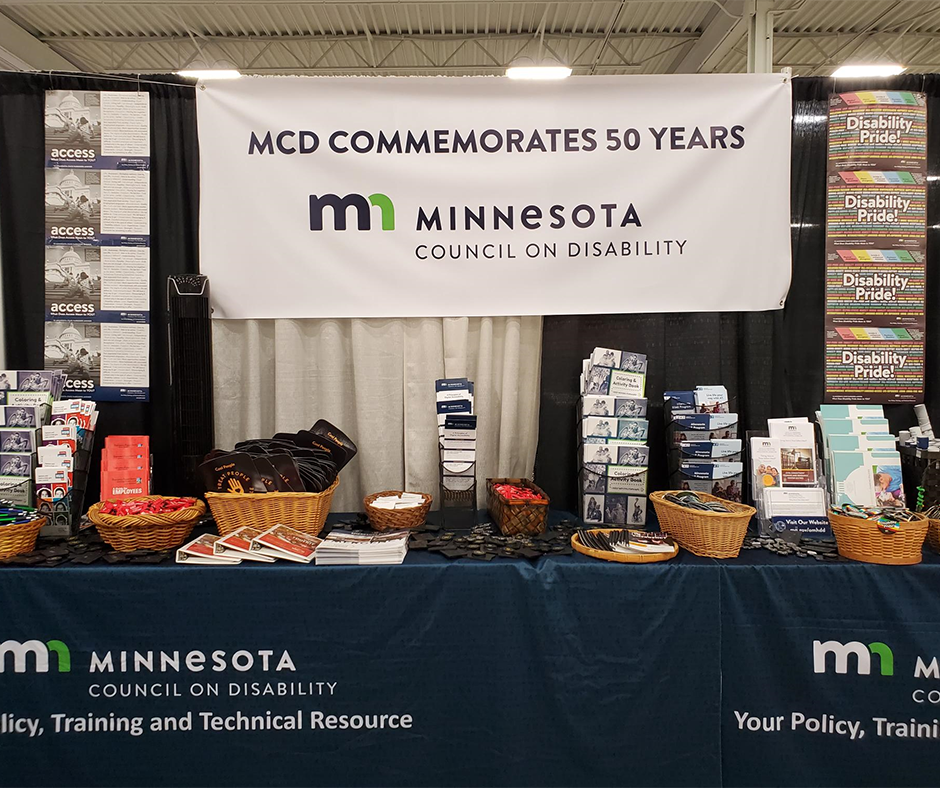 State fair booth. A banner reads, "MCD Commemorates 50 Years. Minnesota Council on Disability." Below it, on a table, are collections of handouts, brochures, buttons, fans, and whistles. 