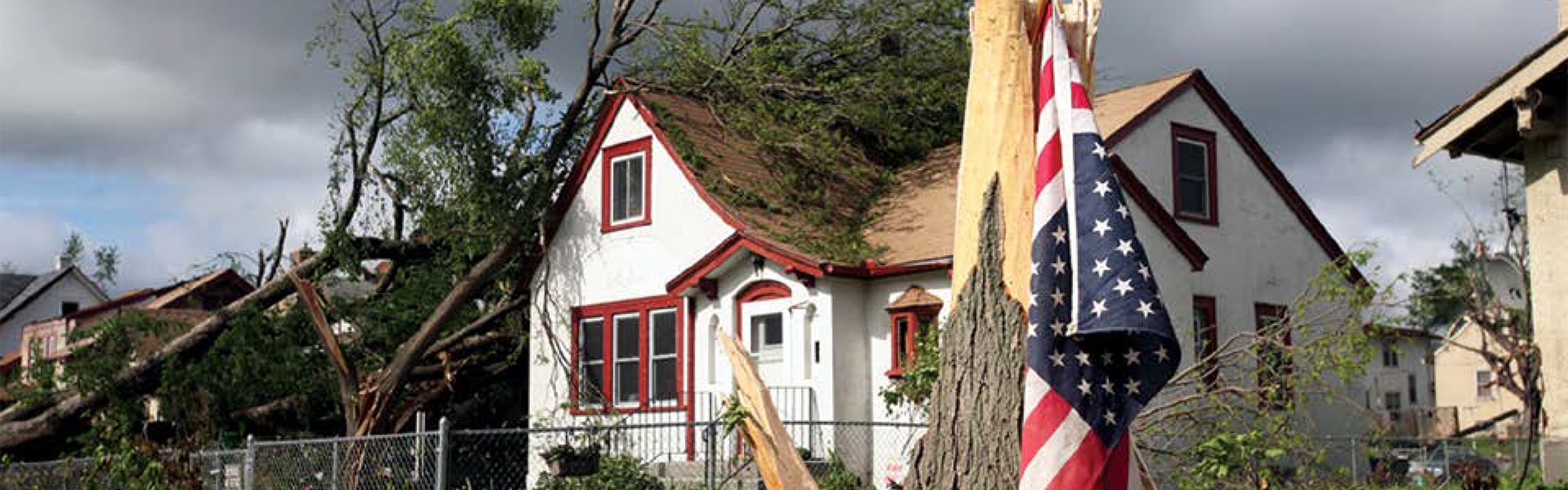 Damaged house surrounded by fallen trees after a storm. A broken tree trunk is in the foreground with an American flag draped over it.