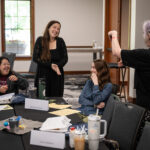 Four woman gathered around a table in a conference room, engaged in discussion. One woman stands and uses sign language, while others smile and laugh. Papers, drinks, and nameplates are visible on the table, including one that reads "MCD Access Consultant."