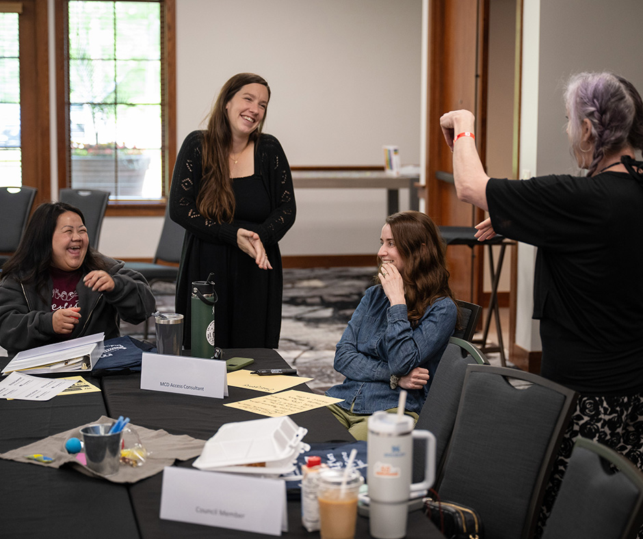 Four woman gathered around a table in a conference room, engaged in discussion. One woman stands and uses sign language, while others smile and laugh. Papers, drinks, and nameplates are visible on the table, including one that reads "MCD Access Consultant."