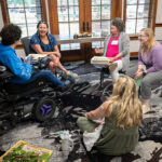 A group of five women sitting together, engaged in conversation. One woman uses a power wheelchair. They are gathered in a room with large windows.