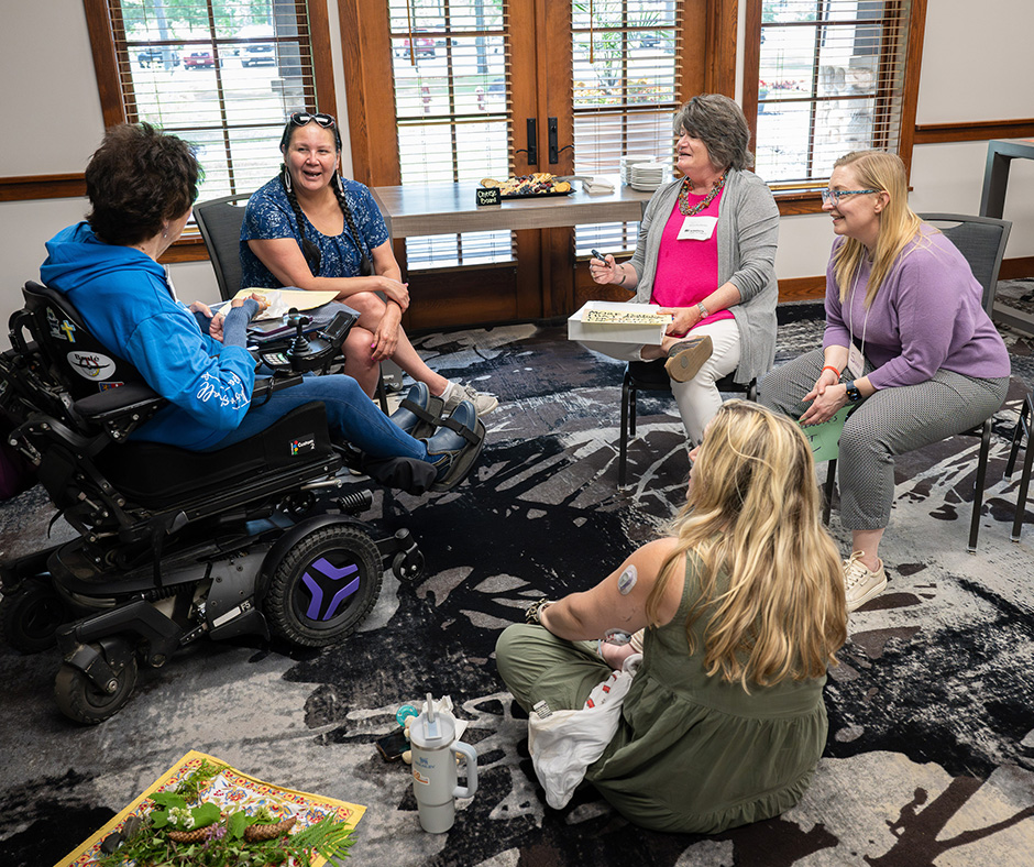 A group of five women sitting together, engaged in conversation. One woman uses a power wheelchair. They are gathered in a room with large windows.