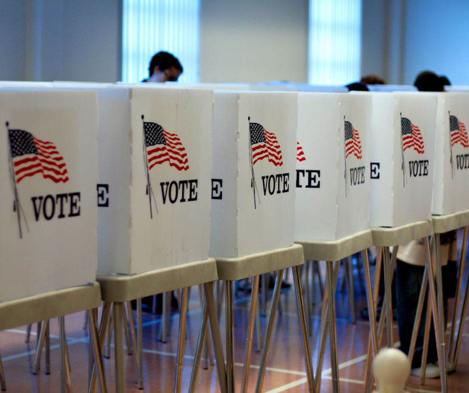 A row of white voting booths with the word 'VOTE' and an American flag printed on the side. People are standing behind the booths, partially visible, inside a large, well-lit room.