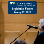 An older woman with gray hair holding a white cane speaks into a microphone at at an event. A banner top center reads: 'Minnesota Council on Disability Legislative Forum, January 27, 2025.' A Minnesota state flag is partially visible in the background.