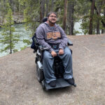 Sumukha Terakanambi, wearing a University of Minnesota hoodie and seated in a power wheelchair, outdoors near a forested area. A river flows behind him, surrounded by pine trees and a dirt path.