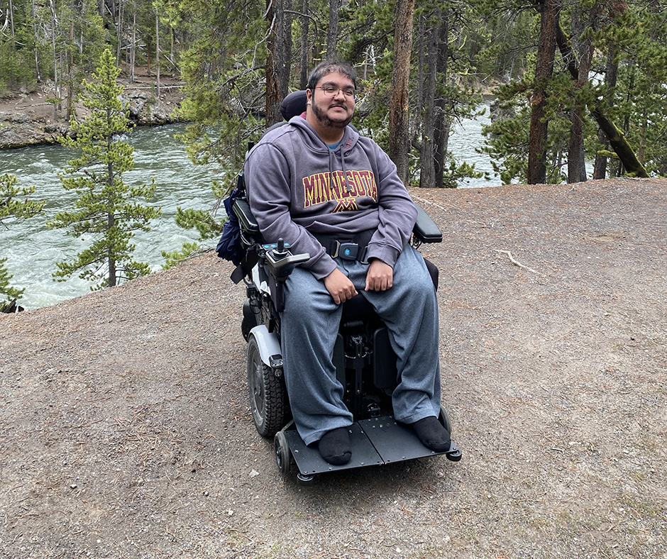 Sumukha Terakanambi, wearing a University of Minnesota hoodie and seated in a power wheelchair, outdoors near a forested area. A river flows behind him, surrounded by pine trees and a dirt path.