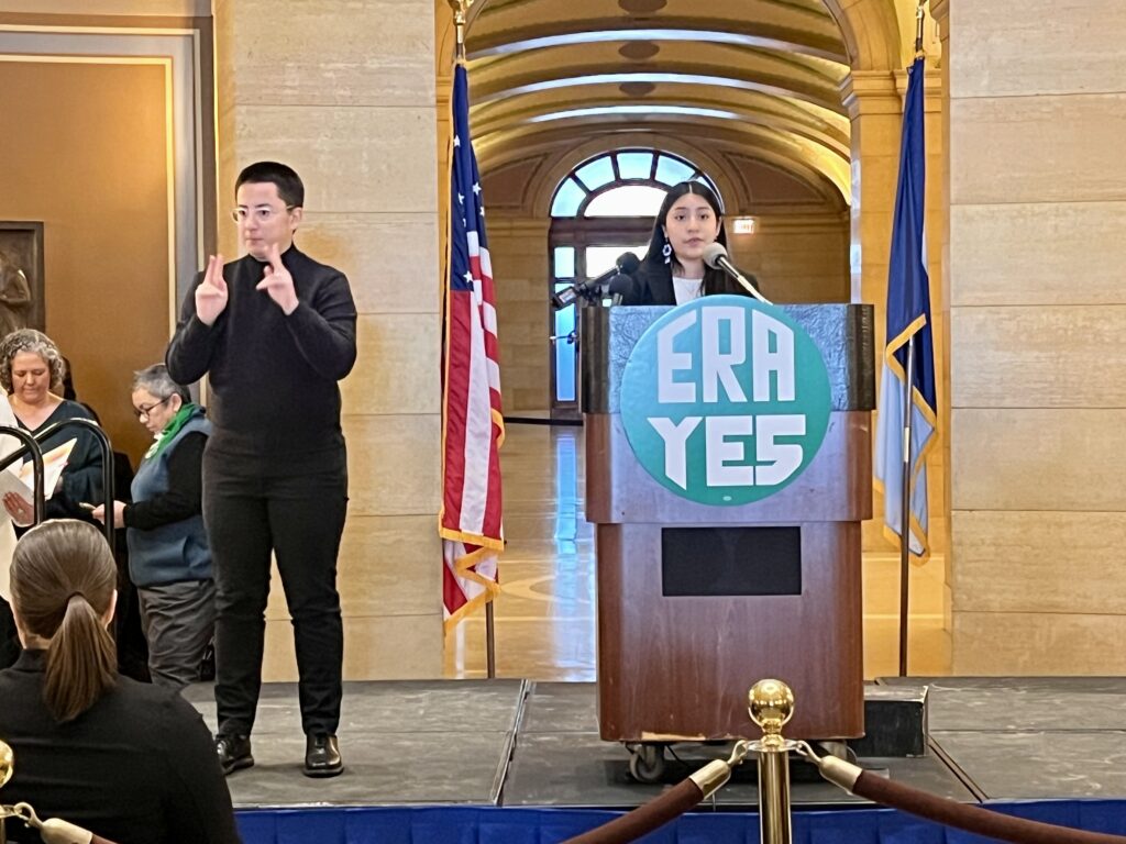 A speaker stands at a podium with a large "ERA YES" sign, inside the Capitol Rotunda. An ASL interpreter signs to the audience. Behind them, an American flag and the Minnesota state flag are displayed.