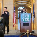 A speaker stands at a podium with a large "ERA YES" sign, inside the Capitol Rotunda. An ASL interpreter signs to the audience. Behind them, an American flag and the Minnesota state flag are displayed.